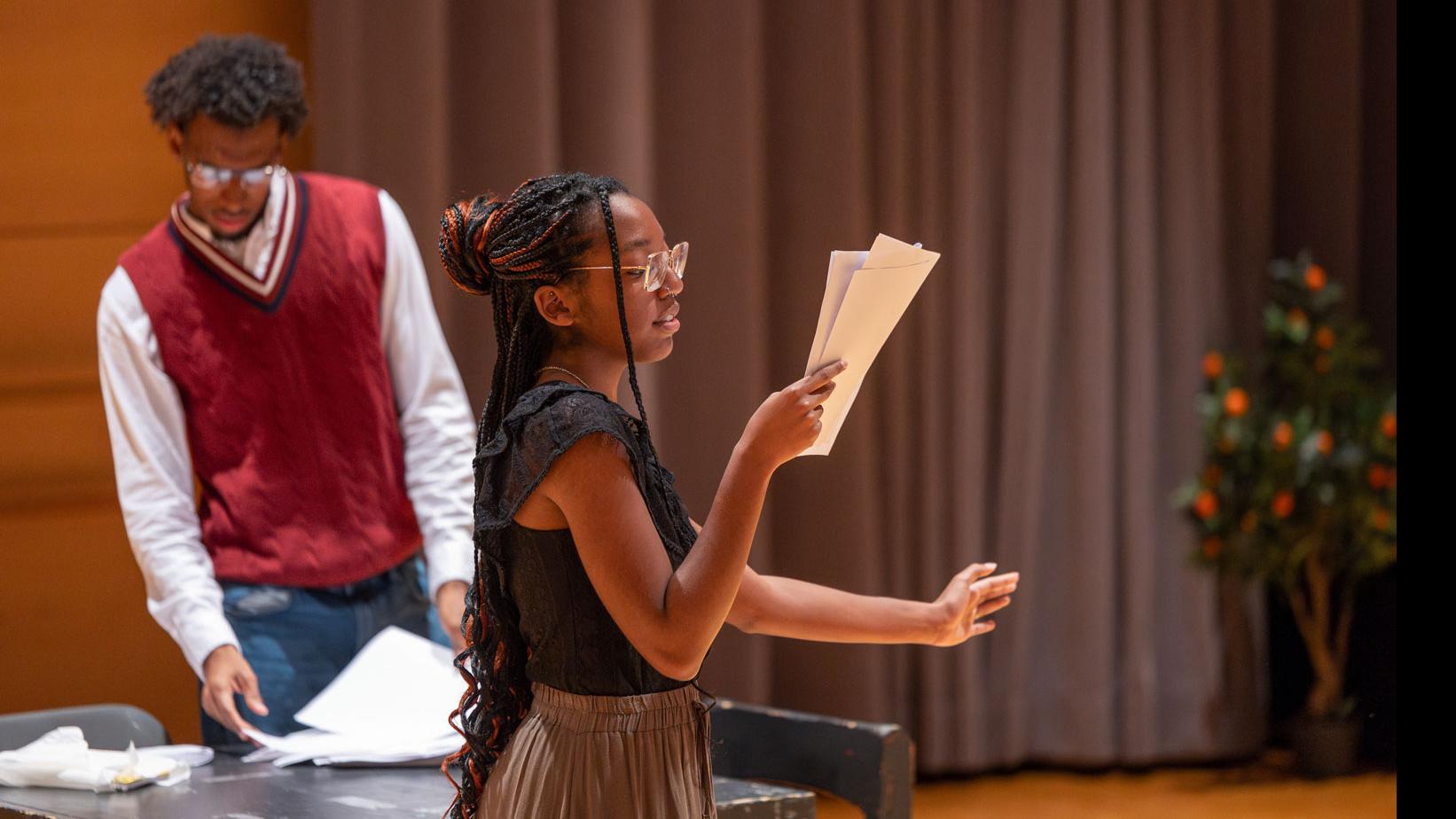 A student stands and looks at a script on stage while another student in the background looks at a script on a table in the Class of ’56 Dance Studio Theatre.