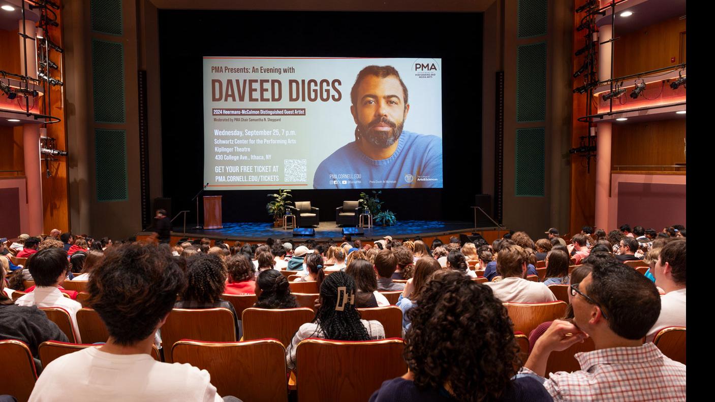 From the back of the orchestra section of the Kiplinger Theatre. In the foreground, a crowd of people sit in the audience, in the background the Kiplinger stage has two chairs, two plants, a podium, and a large screen showing the poster for the event “PMA Presents: An Evening with Daveed Diggs.”