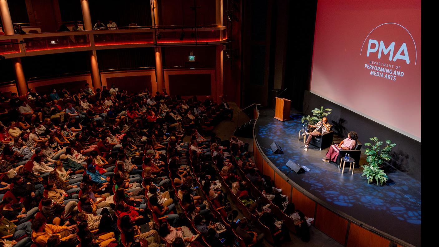 An aerial view from the right side of the orchestra section of the Kiplinger Theatre. A crowd of people sit in the audience, and Daveed Diggs and PMA Department Chair Samantha Noelle Sheppard sit on stage and speak, with a large screen showing the PMA logo.