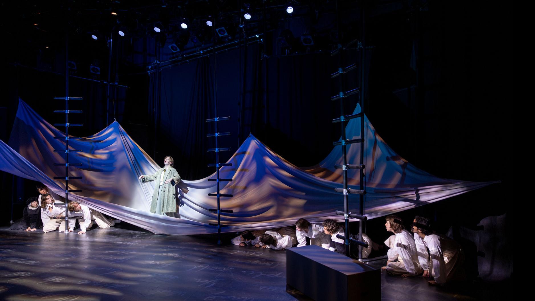 An ensemble of performers spread across the stage, hunched under a sheet made to look like water with one performer spotlit standing on the sheet. 