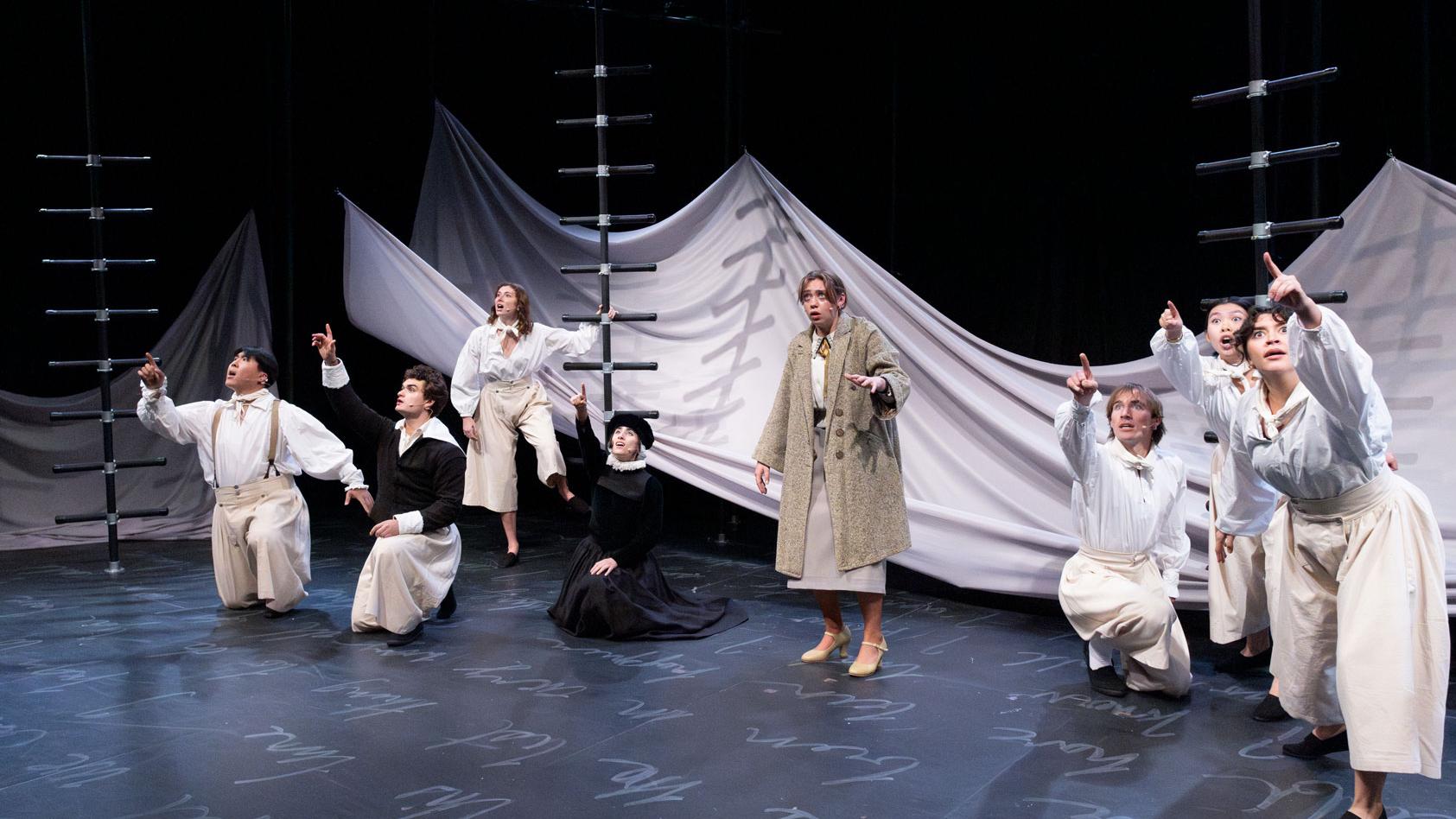 An ensemble of performers pointing outward on a stage set with black ladders, a sheet made to look like water, and white script writing on the ground.