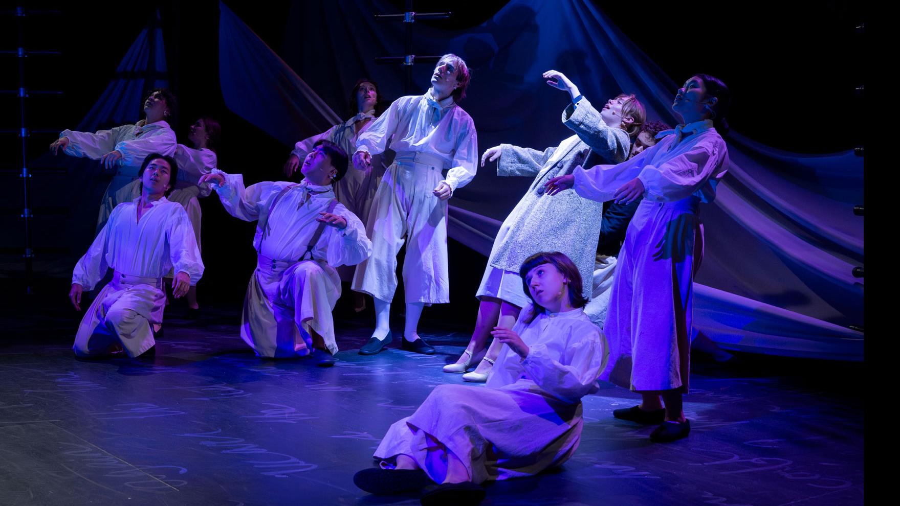 An ensemble of performers sitting and standing on a stage under blue light, with black ladders, a sheet made to look like water, and white script writing on the ground. 