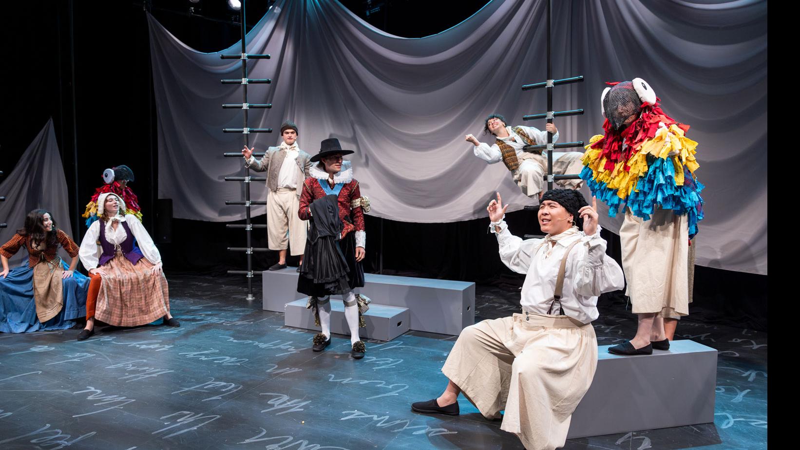 An ensemble of performers scattered across the stage, sitting and standing, on a stage set with black ladders, gray steps and podiums, a sheet made to look like water, and white script writing on the ground. 