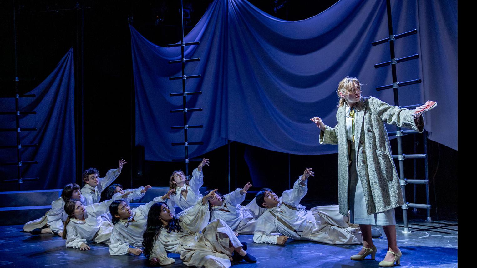 A performer on the right looking off stage, with an ensemble of performers on the ground on the left reaching their arms up, on a set with black ladders, gray steps and podiums, a sheet made to look like water, and white script writing on the ground.