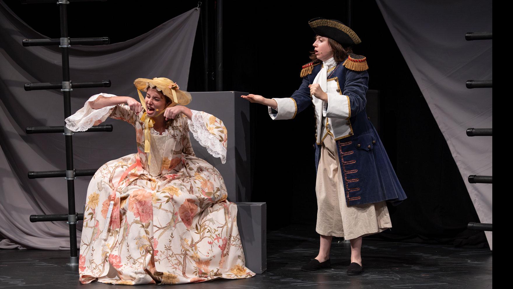 A performer sitting on a gray step on stage with hands in the motion of eating, while another performer reaches out a hand, on a stage set with black ladders and gray steps and podiums. 