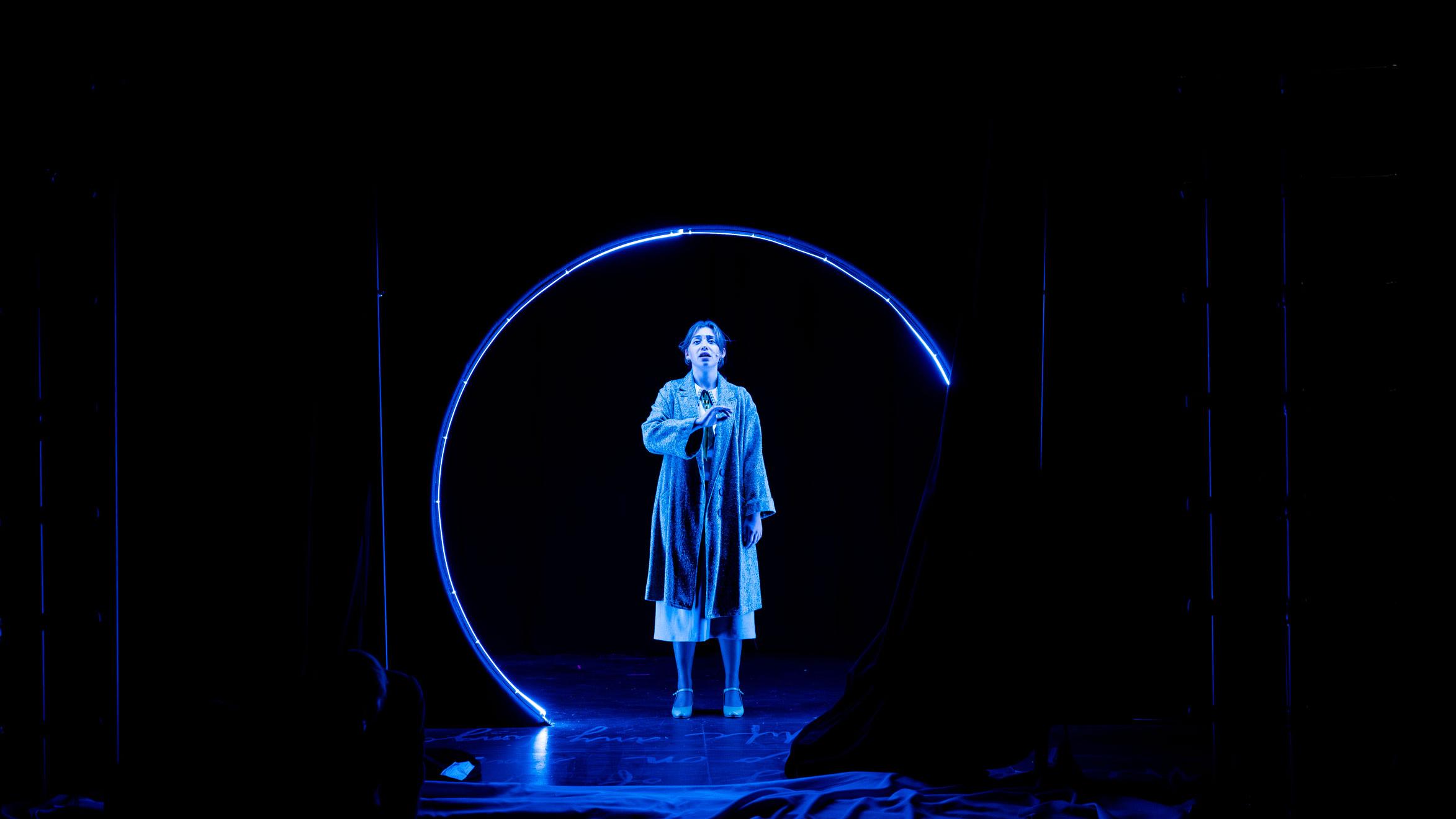 A performer on stage spotlit under blue light looking outward, with a blue circle framing them. 