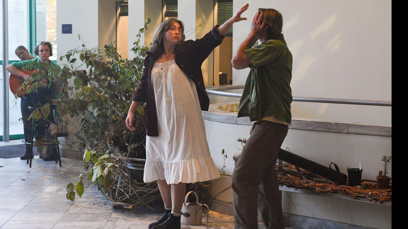 A student performer holds a hand up to another student performer with hands over their face, while a student in the background sits behind potted plants and plays guitar, in the Schwartz Center lobby.