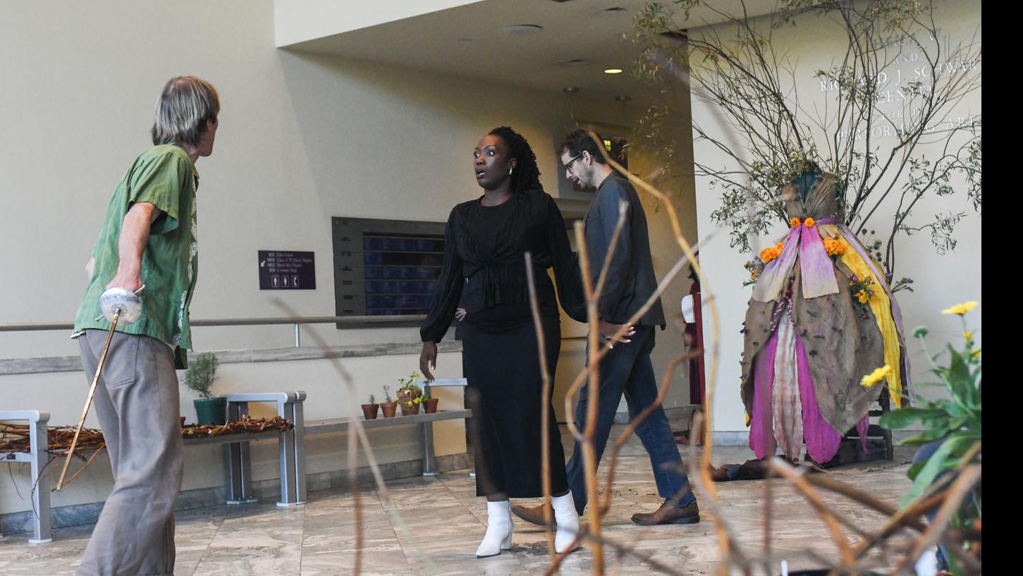 PMA Visiting Lecturer Carolyn Michelle Smith stands facing a person holding a makeshift sword, PMA Senior Lecturer Theo Black steps forward in the background, and a set including a dress with branches coming out of it, potted plants, and dirt on the floor, in the Schwartz Center lobby.