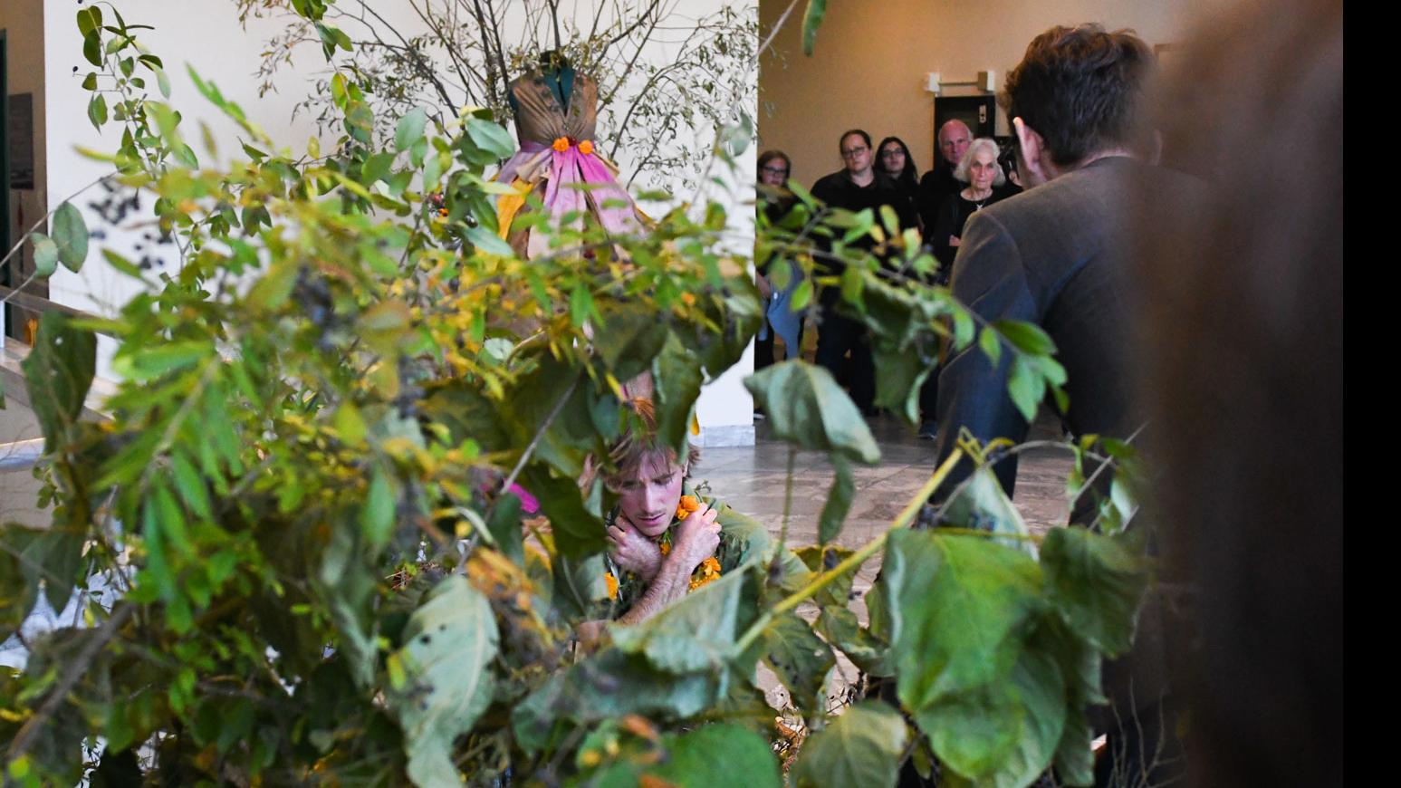 Through plant leaves, a student performer holds their body on the ground, PMA Senior Lecturer Theo Black sits and looks on, while a group of people stand together and watch in the background, on a set including a dress with branches coming out of it in the Schwartz Center lobby.