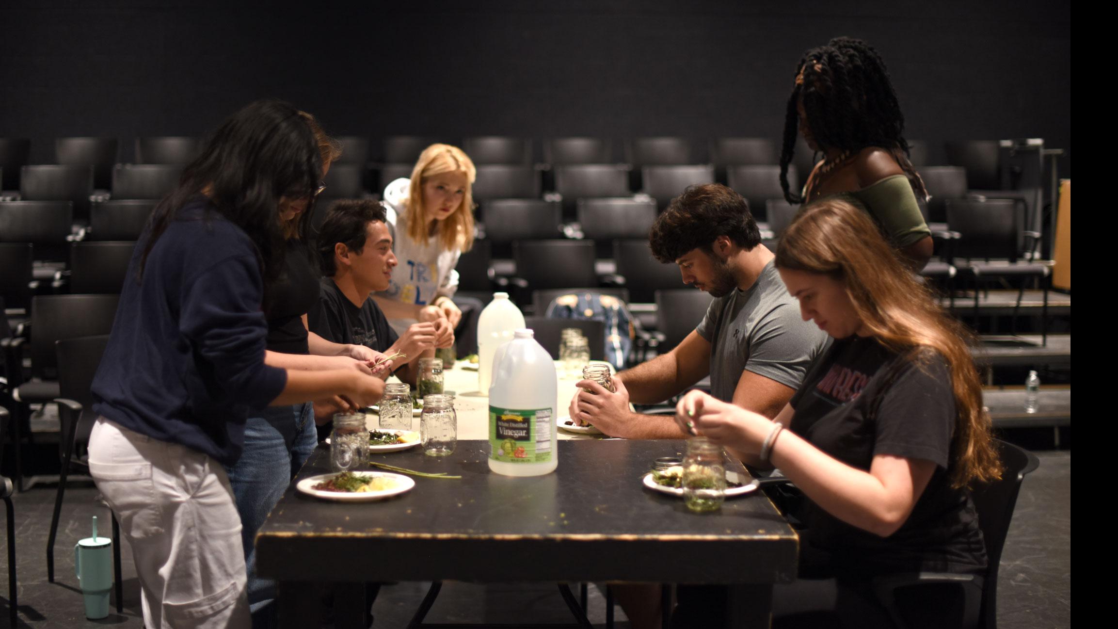 A group of students sit around a table with bottles of vinegar on it, and put spices onto paper plates on stage in the Black Box Theatre. 