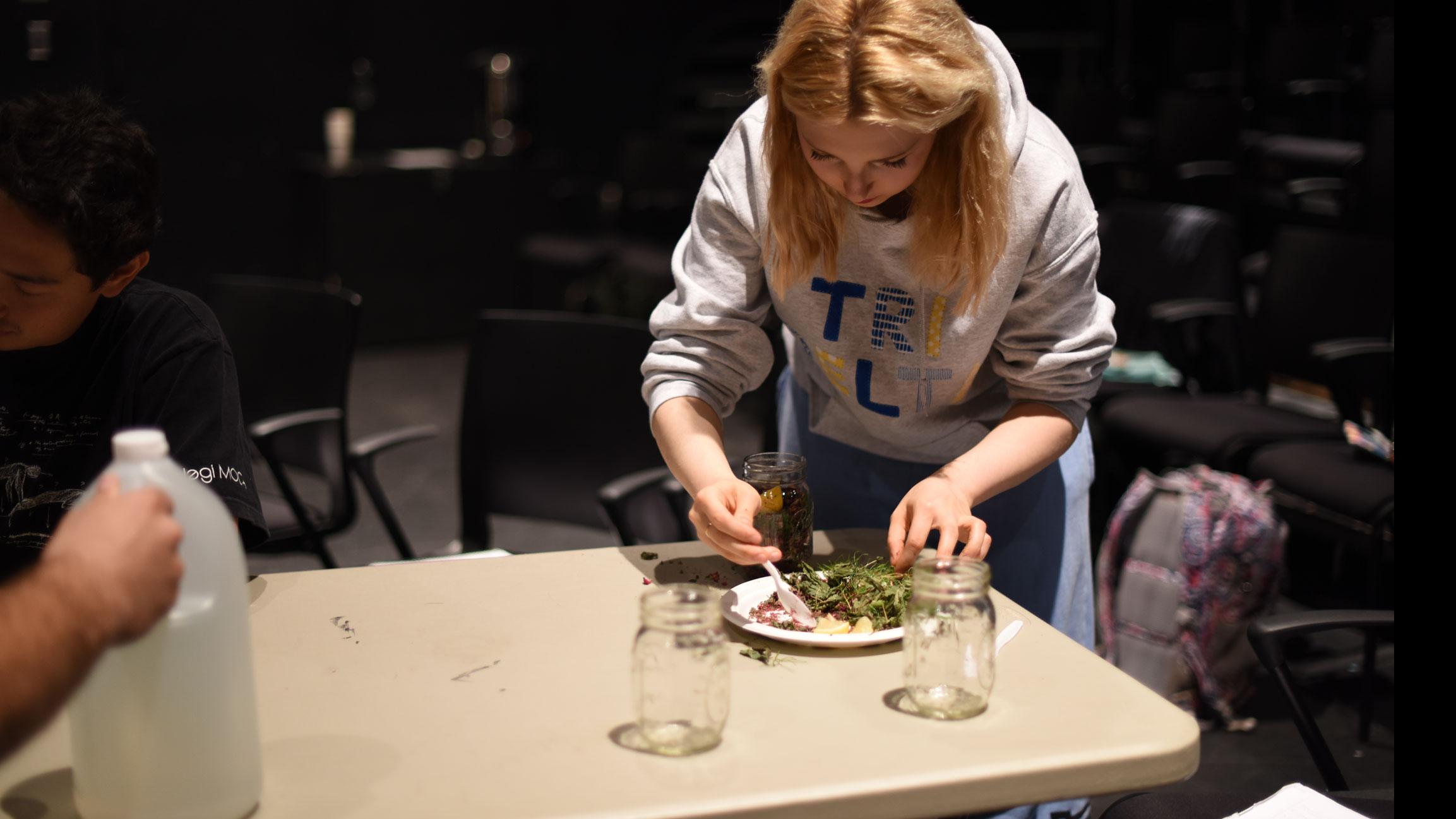 A student stands over a table with a bottle of vinegar on it, and puts spices onto a paper plate on stage in the Black Box Theatre. 