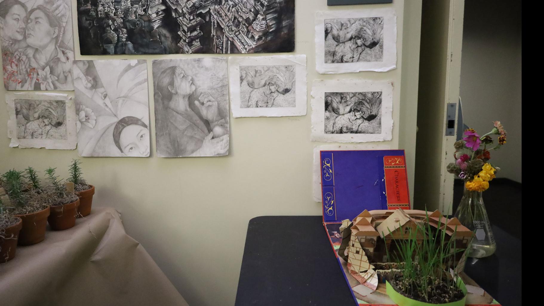 On left, a table of small potted plants, on right a vase of flowers, a plant, and a folded paper house, and in the background, black and white drawings on the wall.