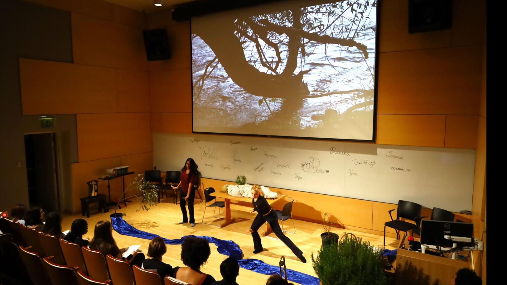 Two students on stage in movement poses next to a long ribbon of blue fabric on the floor, and a big screen showing trees, while a row of students in the audience look on in the Film Forum. 