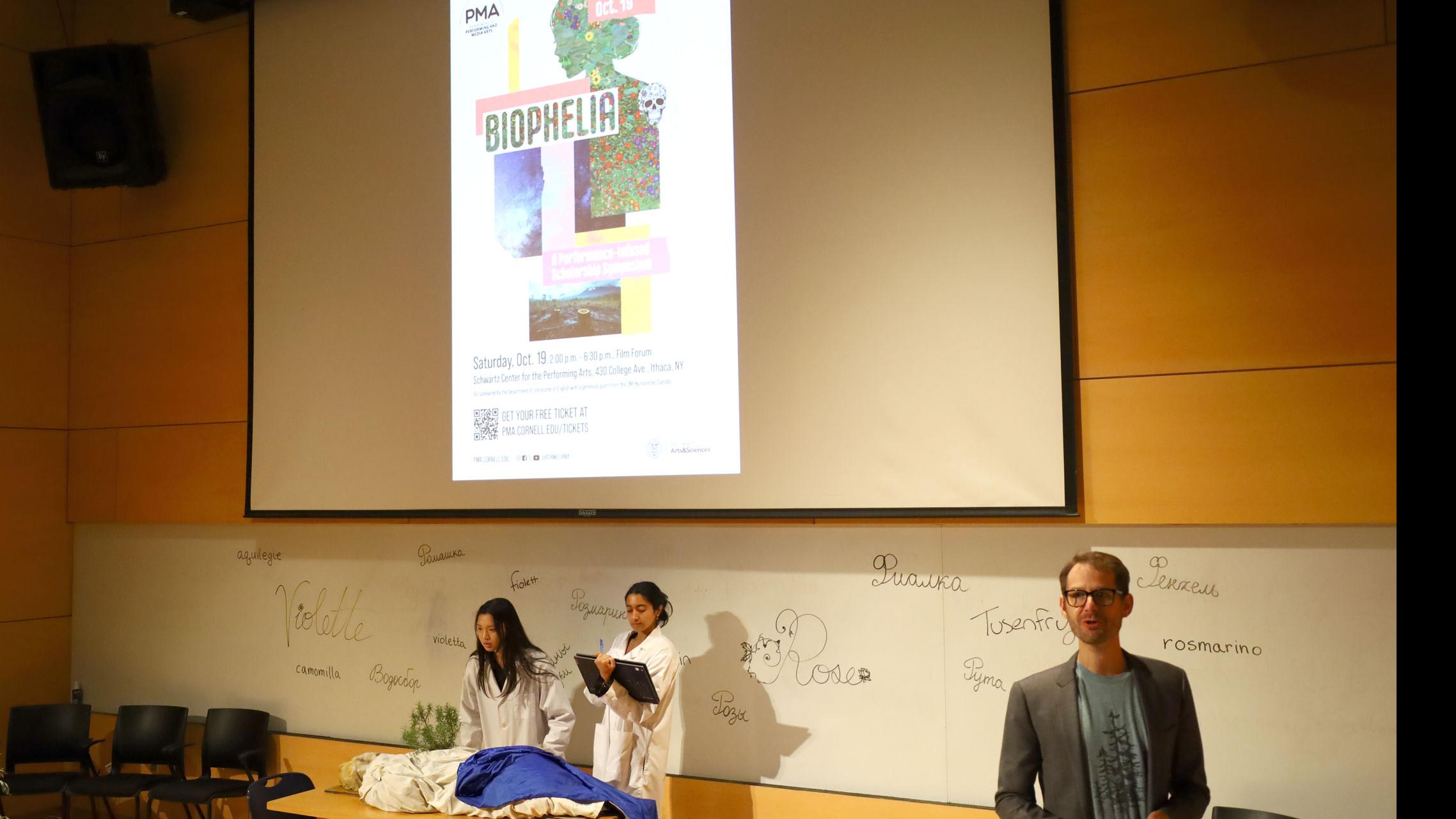 PMA Senior Lecturer Theo Black stands on stage, and two students in white coats look out over a table with blue and white fabric, and a potted plant, against a white board with words for spices written in different languages, and a big screen showing the poster for “BiOphelia,” in the Film Forum.