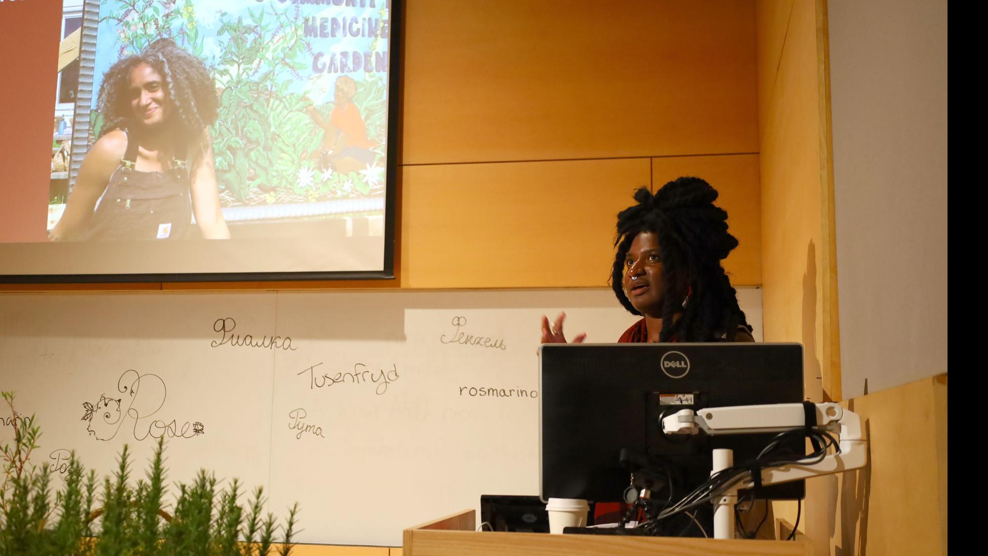 A student on stage at a podium with a computer monitor speaks, against a white board with words for spices written in different languages, and a big screen showing Amanda David, of Rootwork Herbals, in the Film Forum.