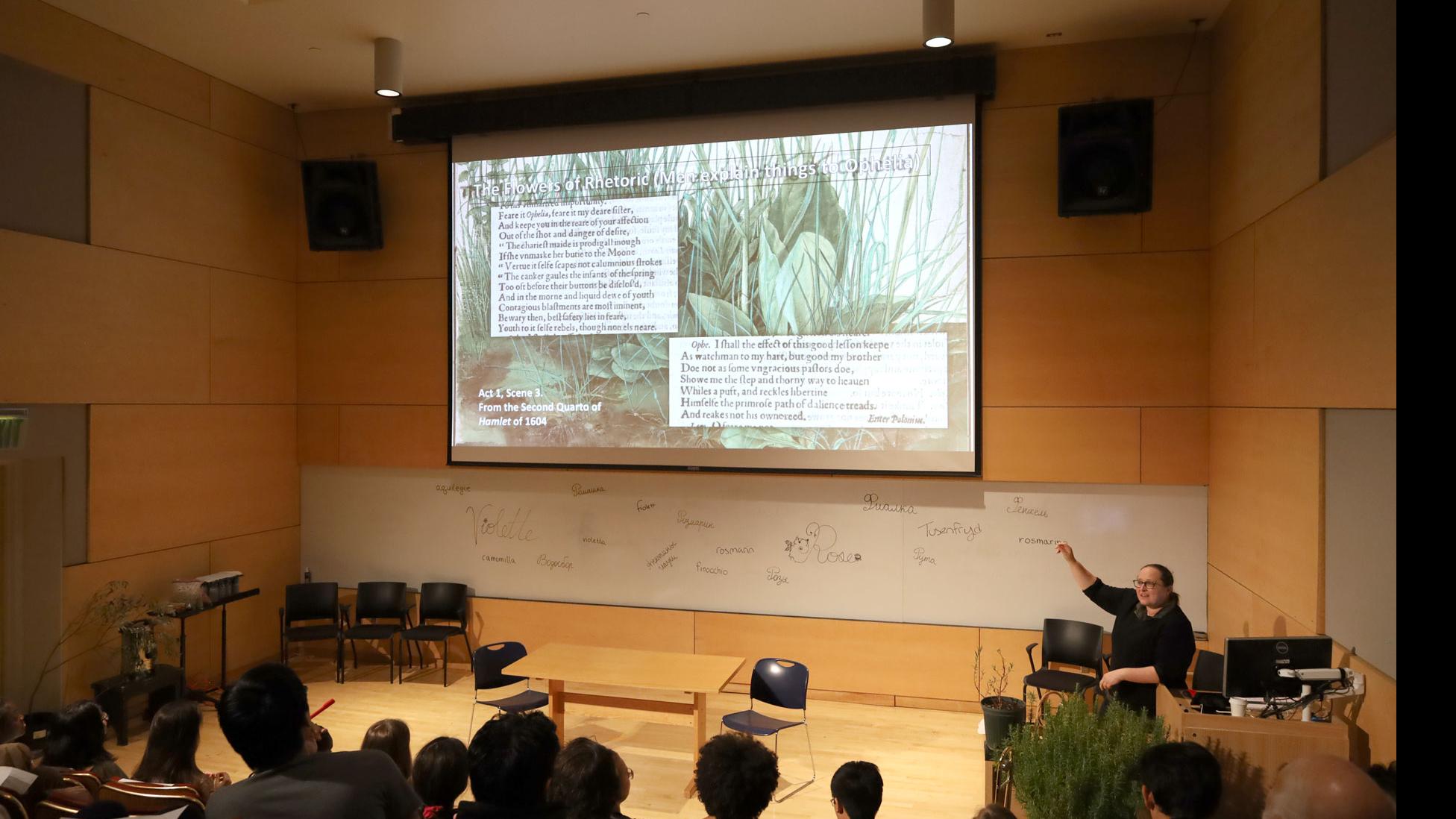 A student on stage at a podium with a computer monitor speaks, against a white board with words for spices written in different languages, and a big screen showing “The Flowers of Rhetoric” in the Film Forum.