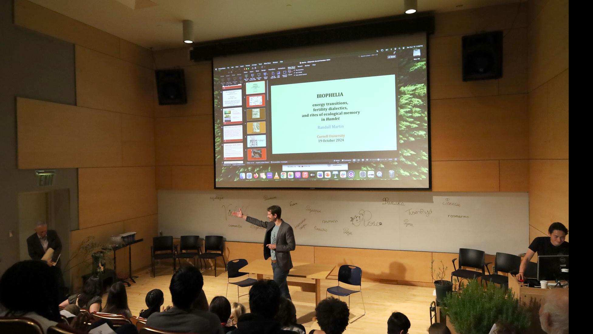 PMA Senior Lecturer Theo Black on stage extends an arm toward a person entering a door from the left, against a white board with words for spices written in different languages, and a big screen showing a slide with the text “Biophelia” and “Randall Martin” in the Film Forum