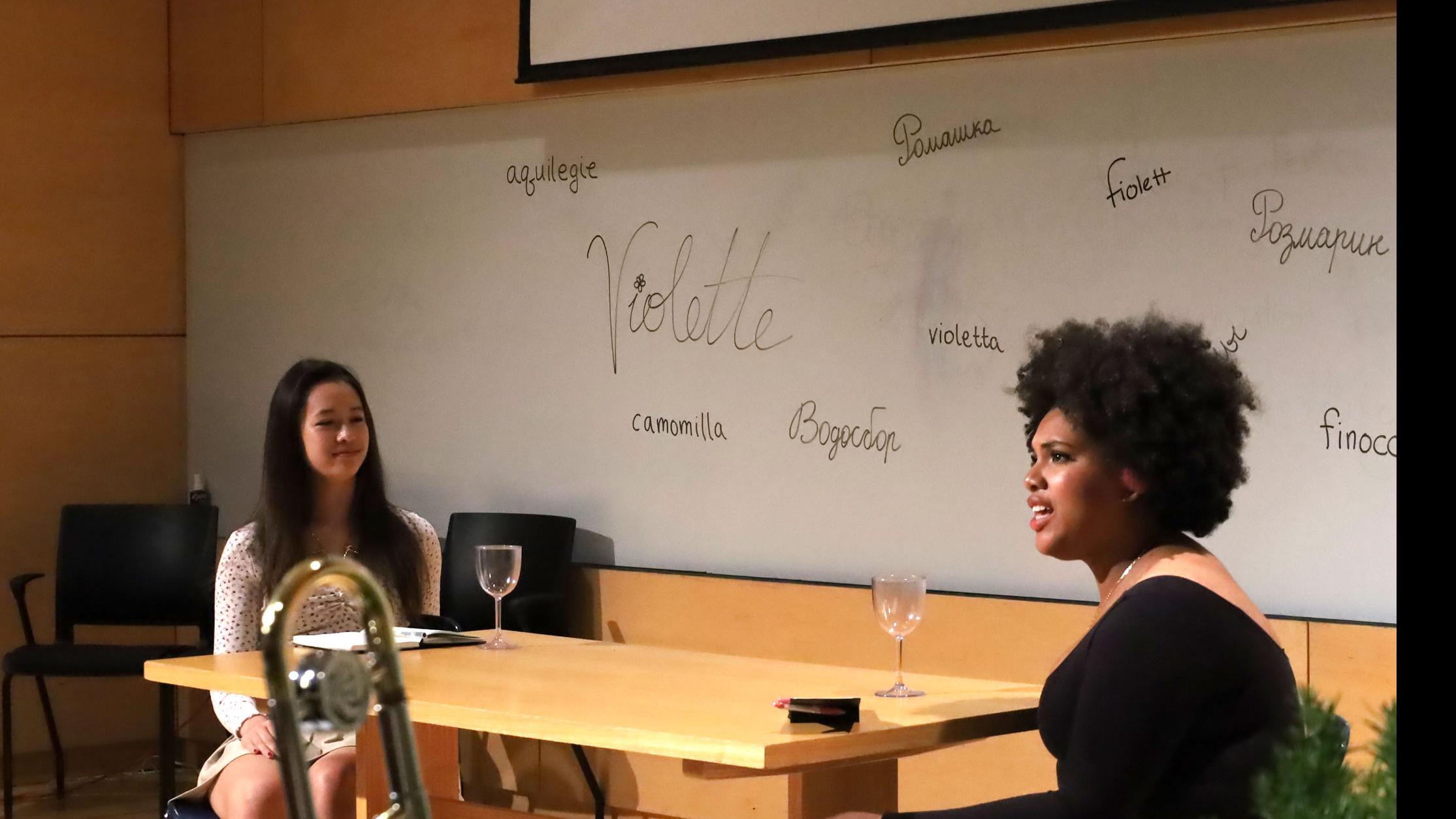 Two students on stage sit at a table with two wine glasses and speak, against a white board with words for spices written in different languages, in the Film Forum.