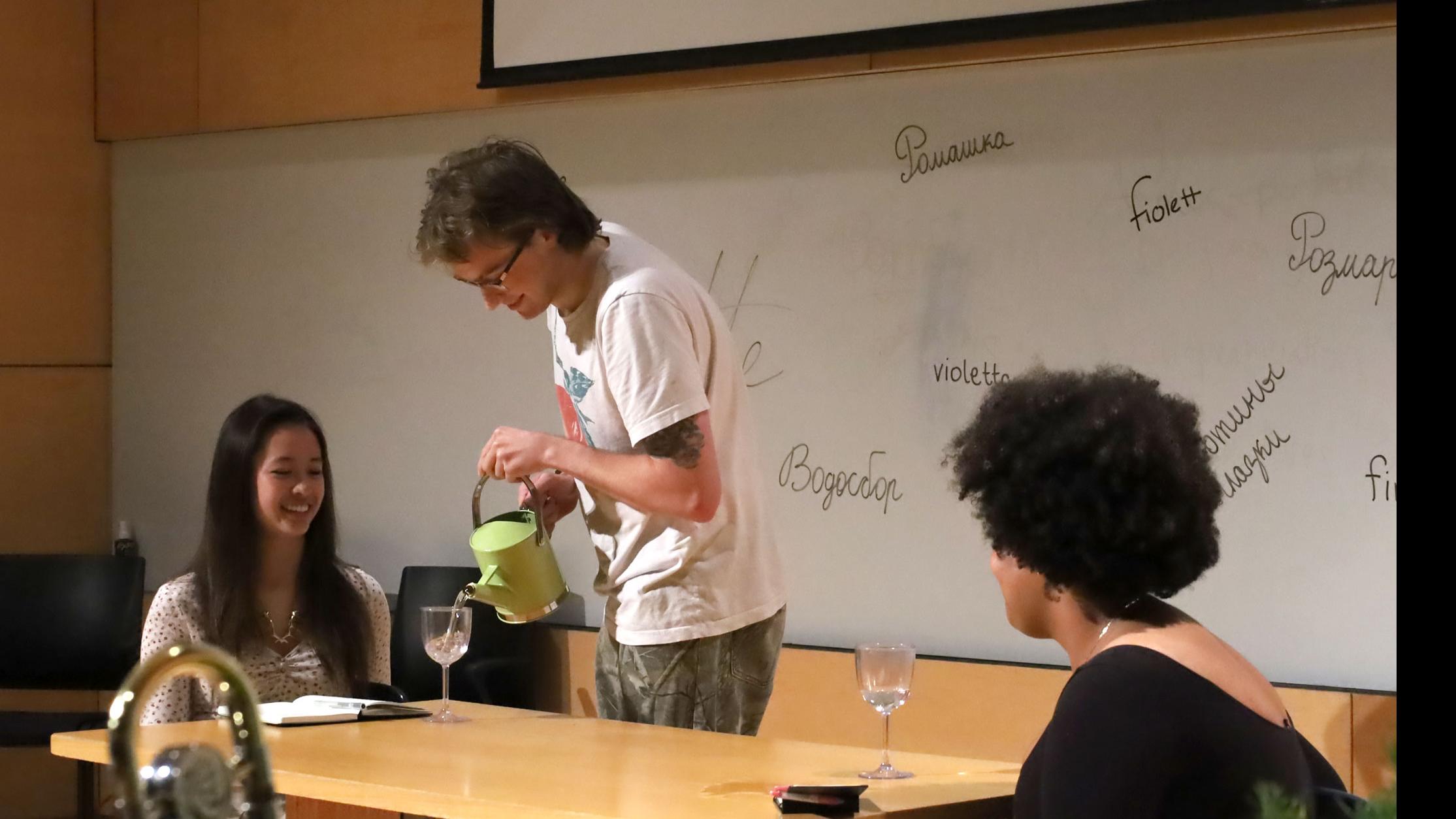 Two students on stage sit at a table with two wine glasses, while another student pours water into one of the glasses, against a white board with words for spices written in different languages, in the Film Forum.