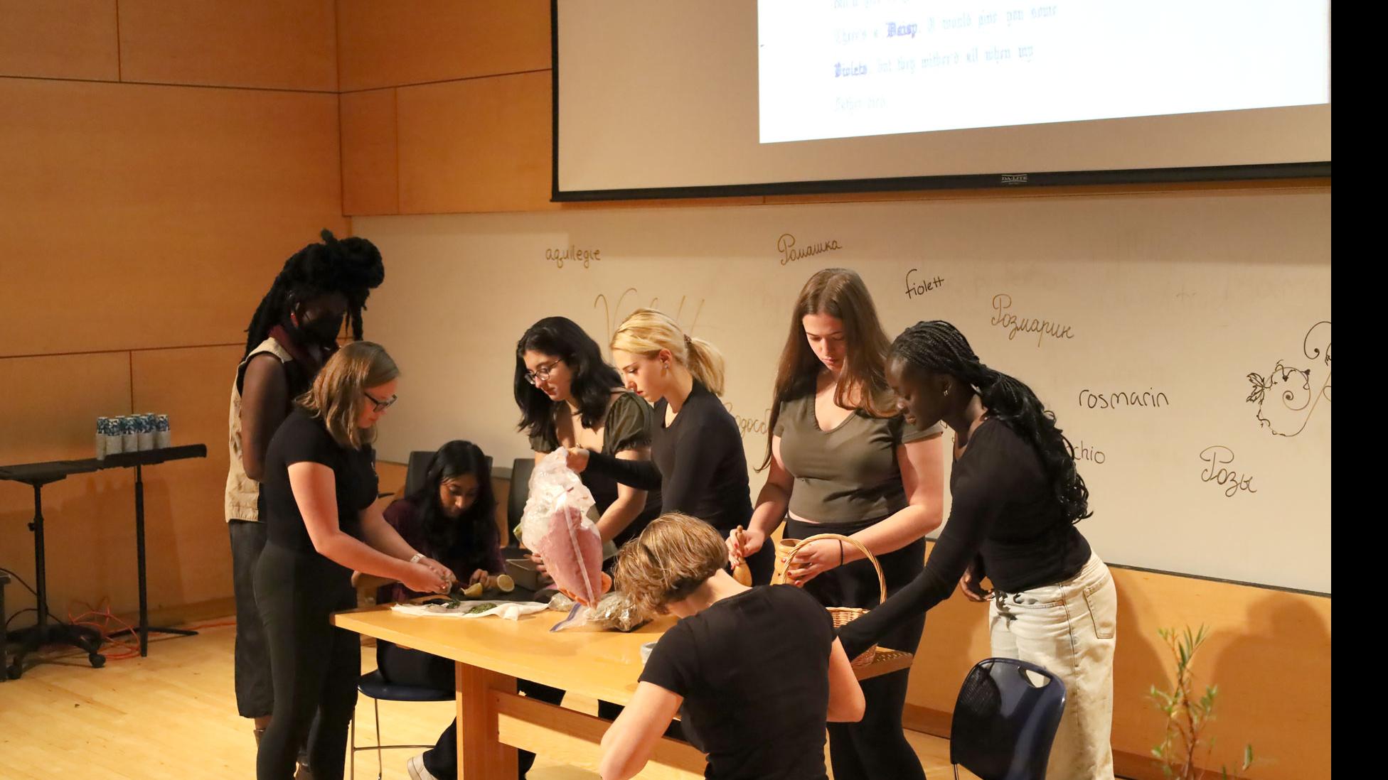 A group of students on stage stand and sit at a table looking down at various plants, against a white board with words for spices written in different languages, in the Film Forum.