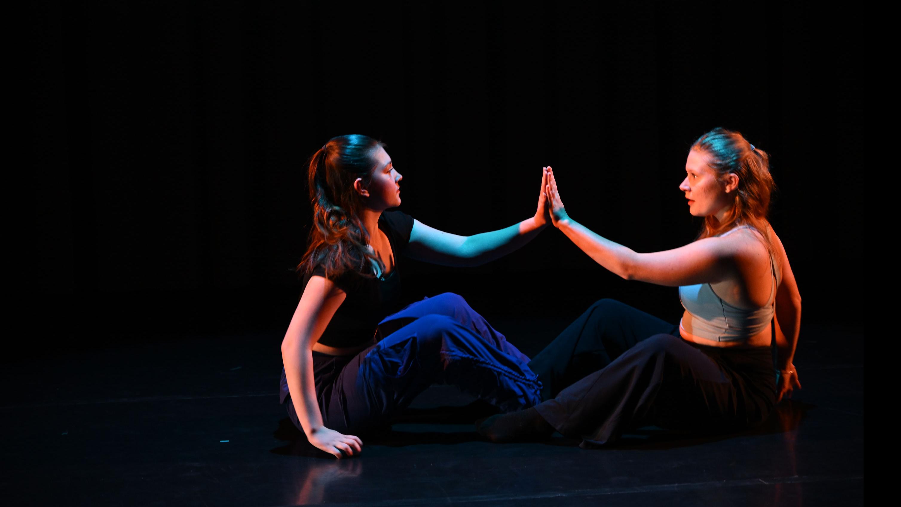 Two performers on stage sit on the ground with a hand each pressed together, in the Class of ’56 Dance Studio Theatre.