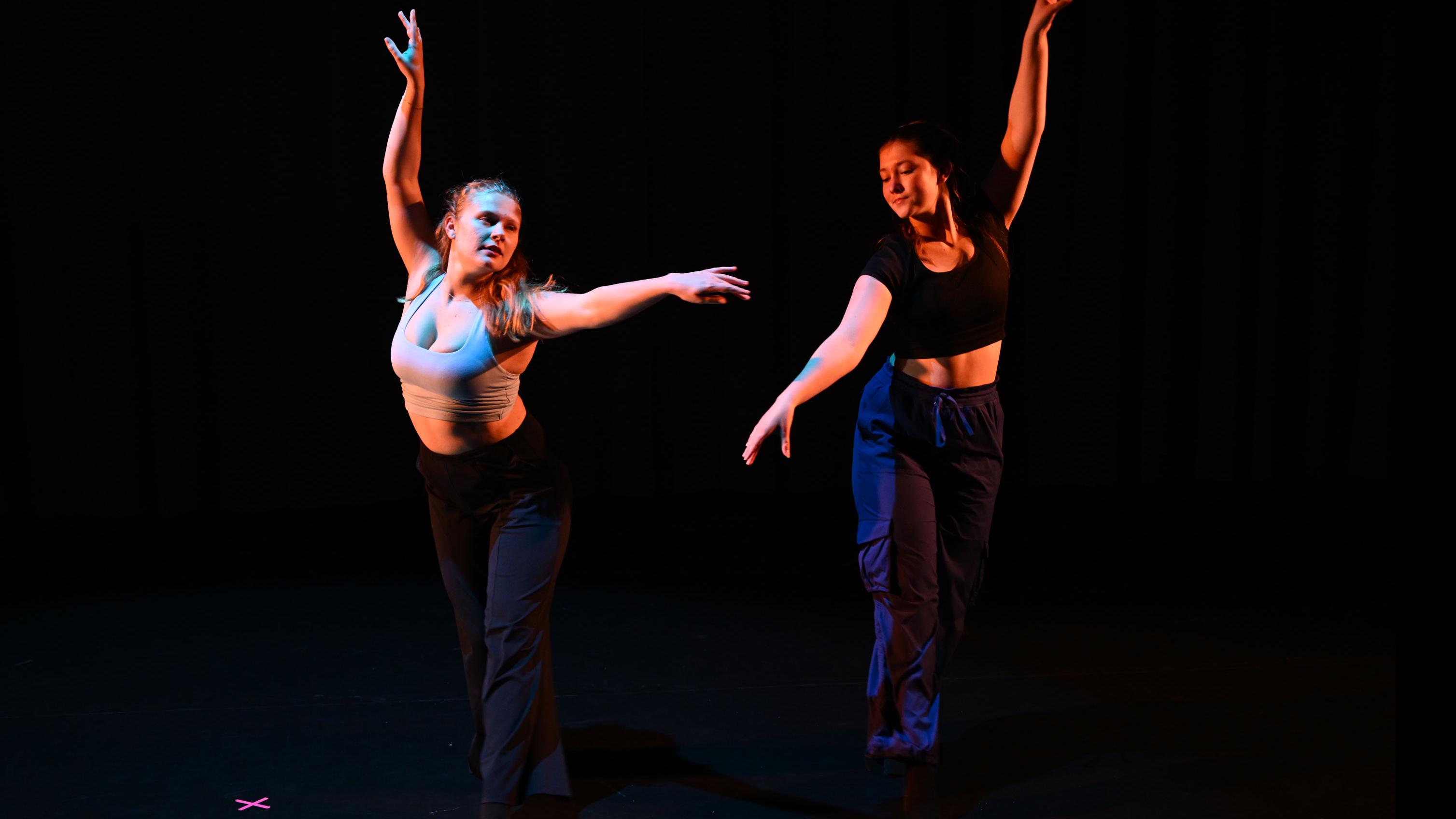 Two performers on stage in mirrored movement poses, with one foot forward and one arm raised, in the Class of ’56 Dance Studio Theatre