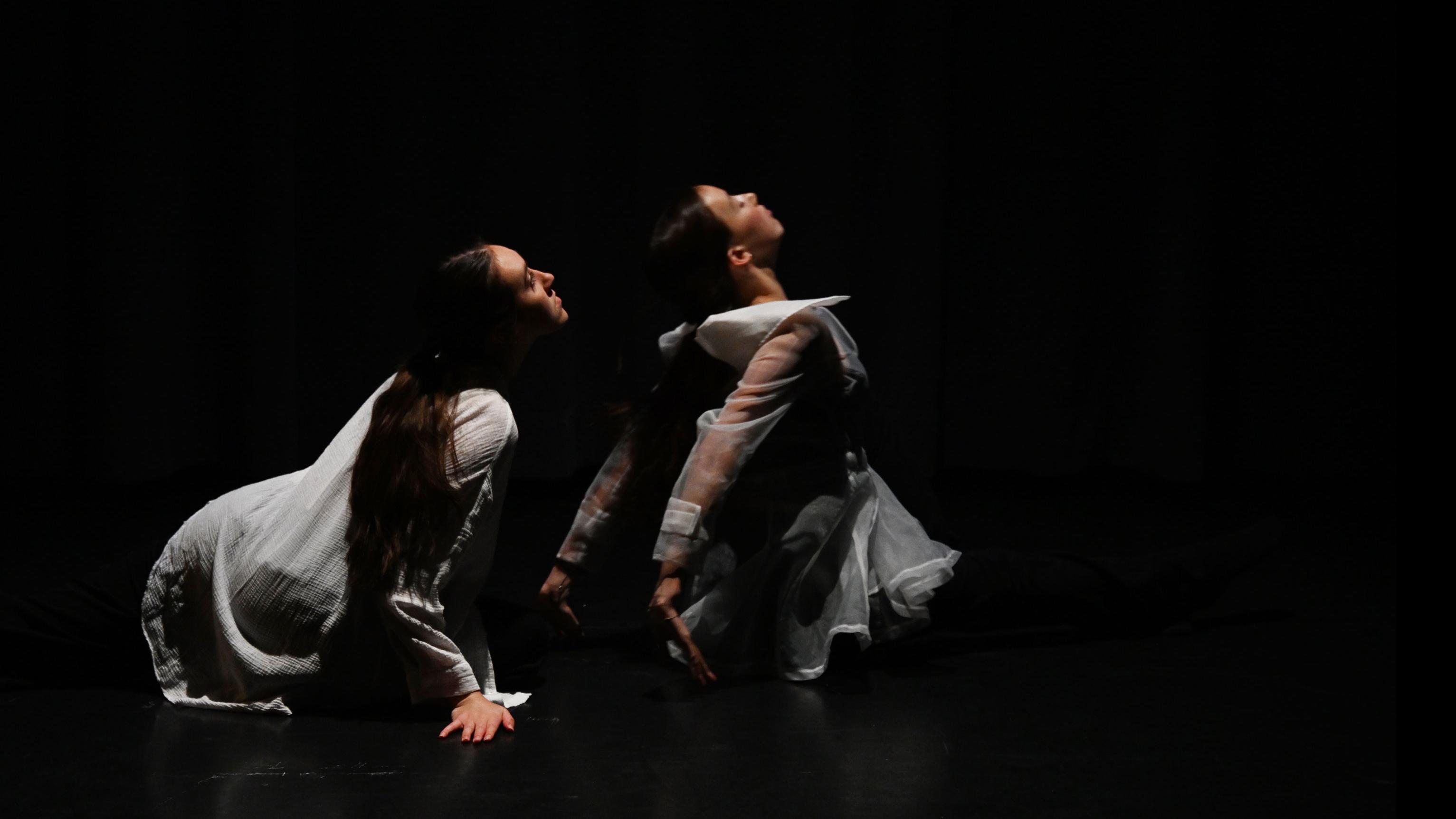 Two performers on stage on the ground tilt their heads up in the same direction, in the Class of ’56 Dance Studio Theatre.