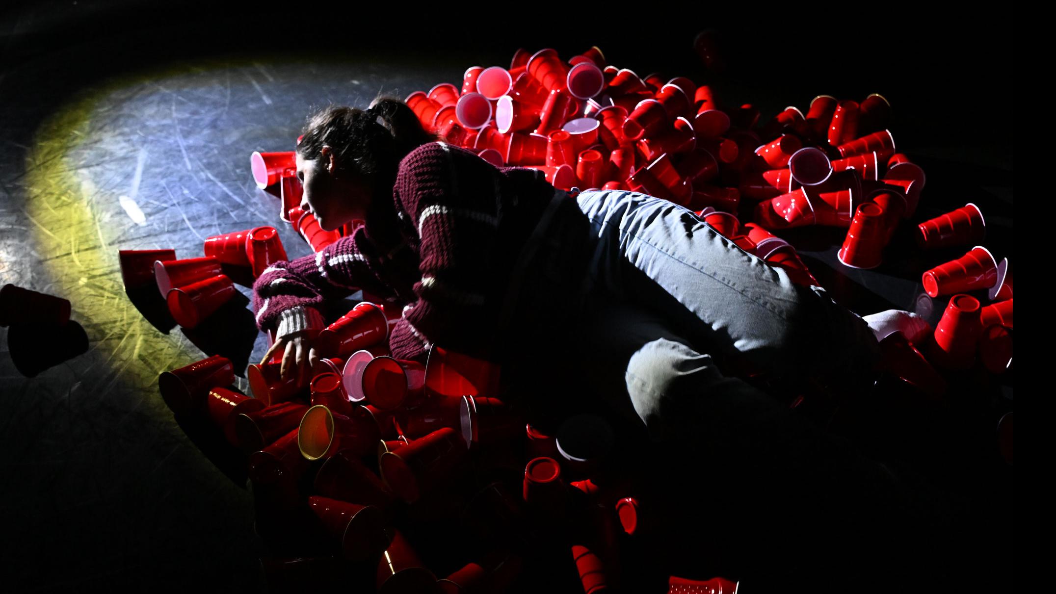 A performer on stage lies in a pile of red solo cups under a spotlight, in the Class of ’56 Dance Studio Theatre.