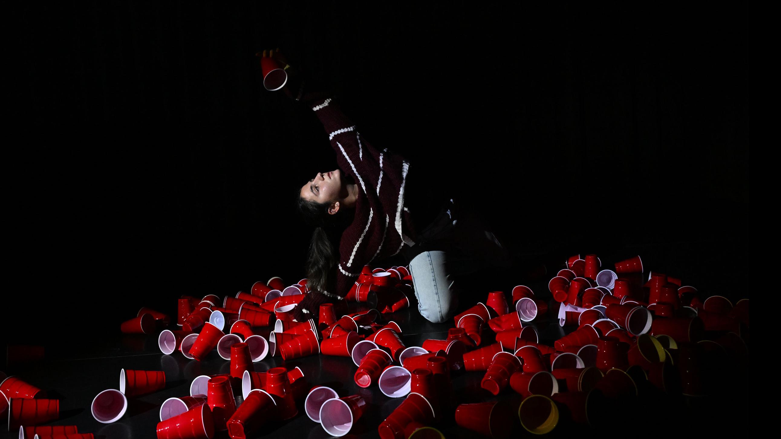 A performer on stage on the ground raises an arm up and holds a red solo cop, amongst a pile of red solo cups on the ground, in the Class of ’56 Dance Studio Theatre.