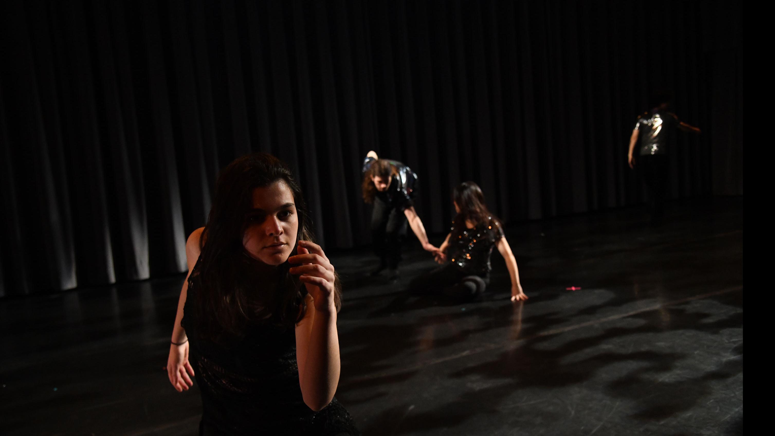 In the foreground, a performer on stage sits on the ground and reaches forward, and in the background, three performers sit and stand in various movement poses, in the Class of’56 Dance Studio Theatre.