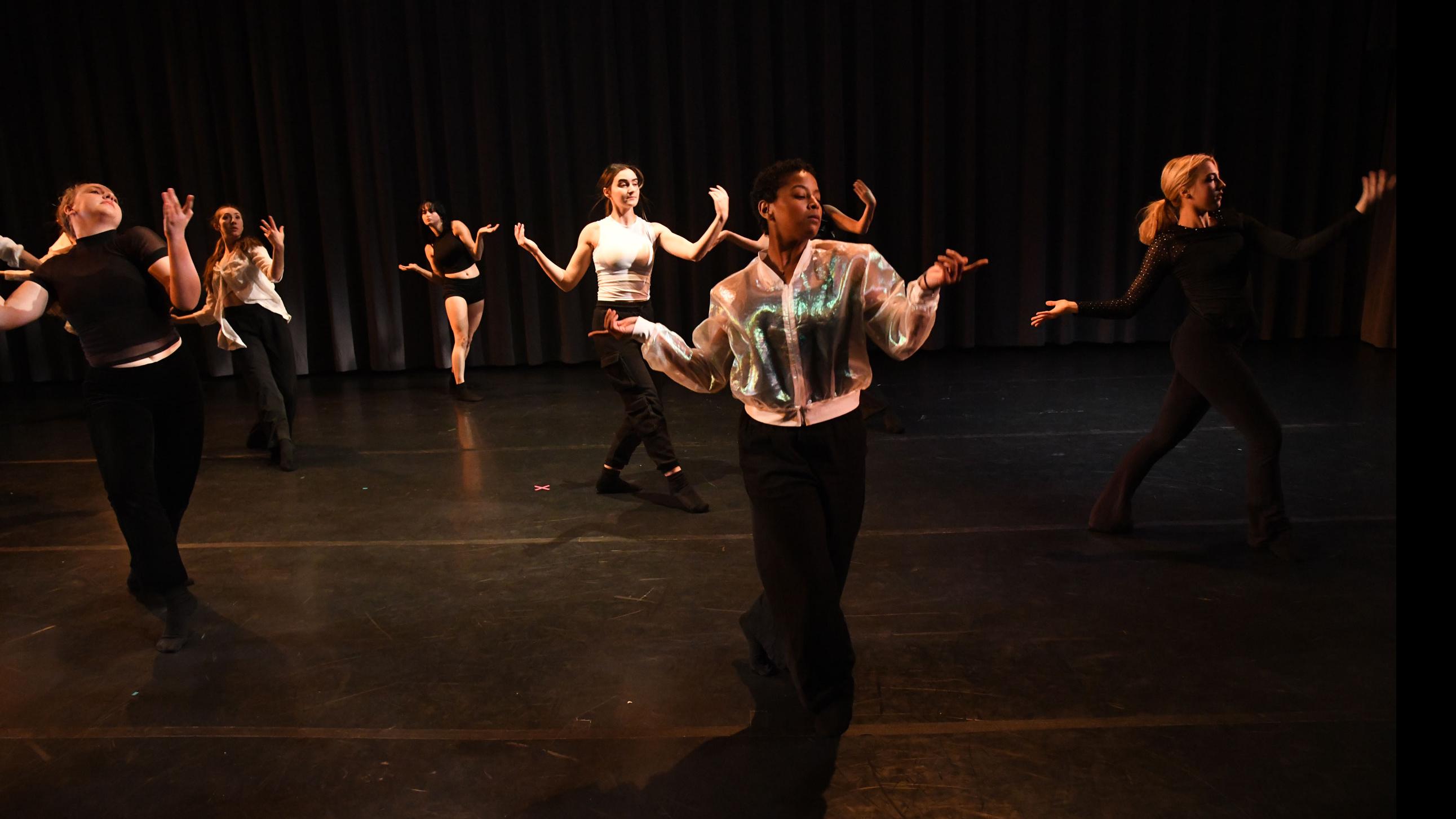 An ensemble of performers spread across the stage in similar movement poses bend their arms out to the side with one leg crossed in front of the other, in the Class of ’56 Dance Studio Theatre.