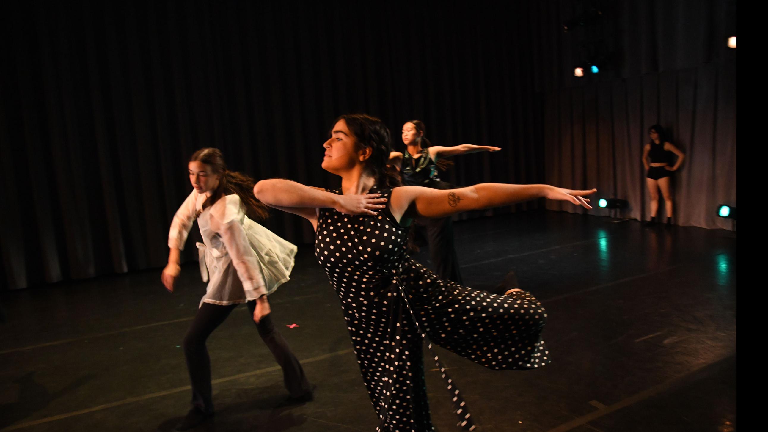 In the foreground, three performers on stage in a triangle step forward and reach an arm back, while one performer in the background stands by a curtain, in the Class of ’56 Dance Studio Theatre.