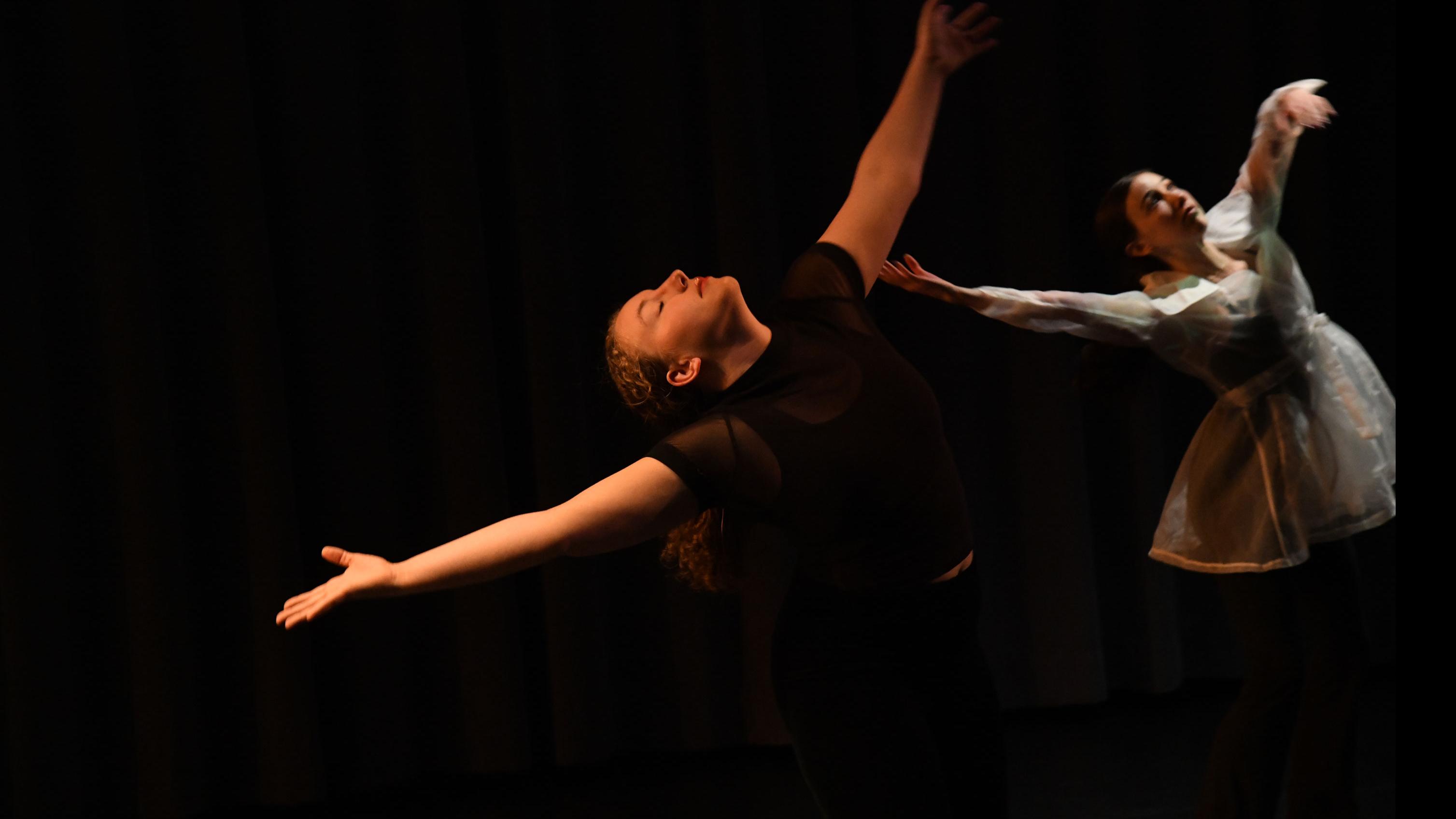 Two performers on stage in similar movement poses with arms out and faces tilted up, in the Class of ’56 Dance Studio Theatre.