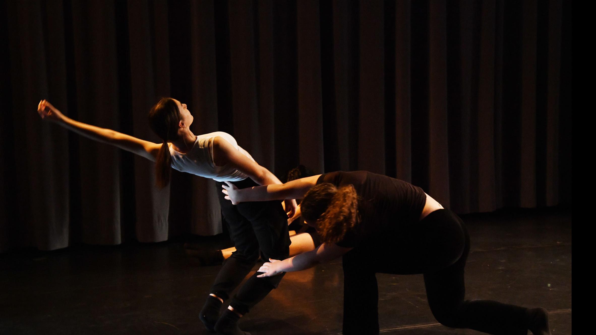 A performer on stage bends forward and holds up another performer who leans backwards with an arm arced up, in the Class of ’56 Dance Studio Theatre.