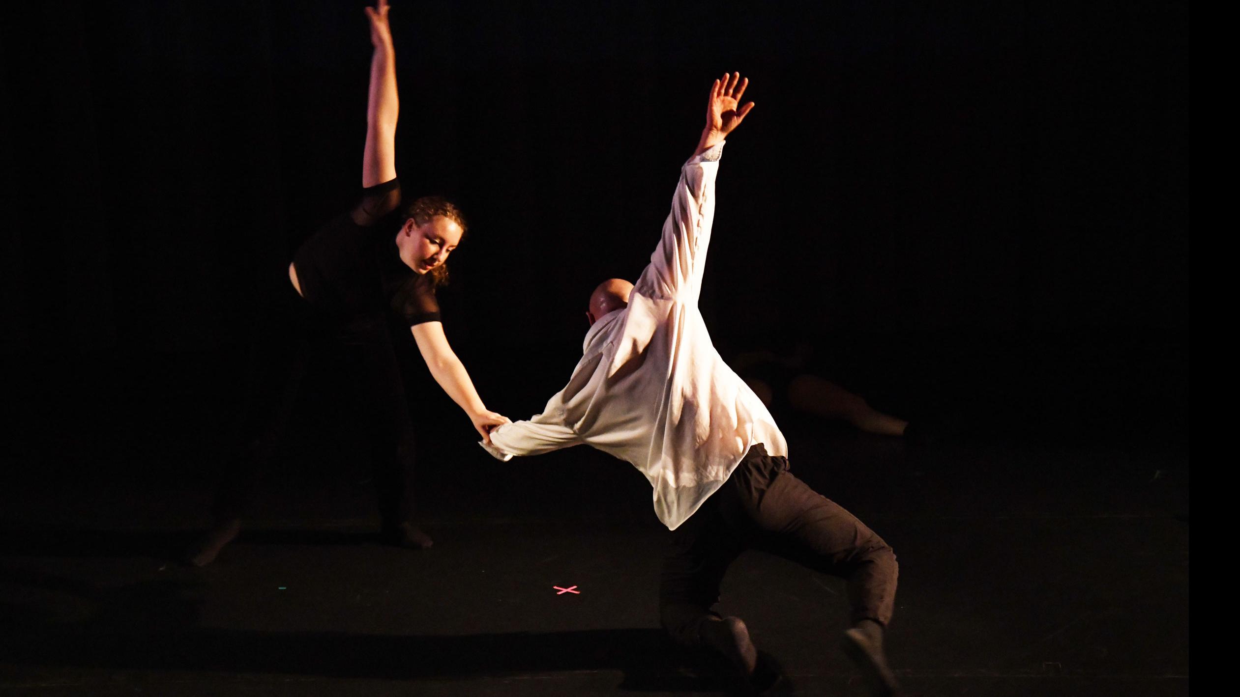 Two performers on stage in mirrored movement poses hold each other by a hand, and raise an arm behind them, in the Class of ’56 Dance Studio Theatre.