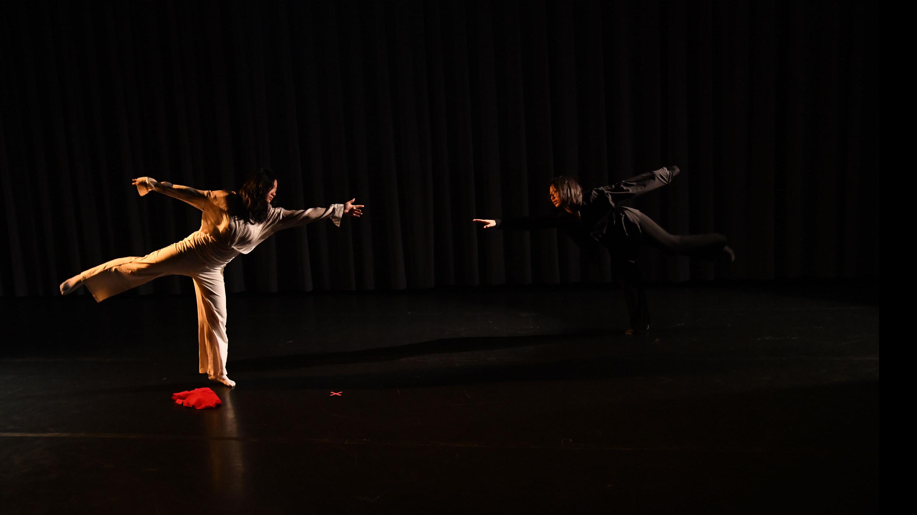 Two performers on stage in mirrored movement poses balance on one foot with one foot back and reach towards the other performer, in the Class of ’56 Dance Studio Theatre.