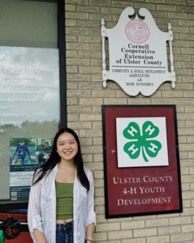 Kailey Hu Ye standing in front of Cornell Cooperative Extension of Ulster County building by the Ulster County 4-H Youth Development sign