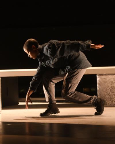 Student dancer Taylor Janeen Pryor stepping forward with her back leg bent, reaching down towards the floor on a stage against a black background.   Image by Thomas Hoebbel Photography, featuring Taylor Janeen Pryor in “This Table Has Been a House in the Rain.”